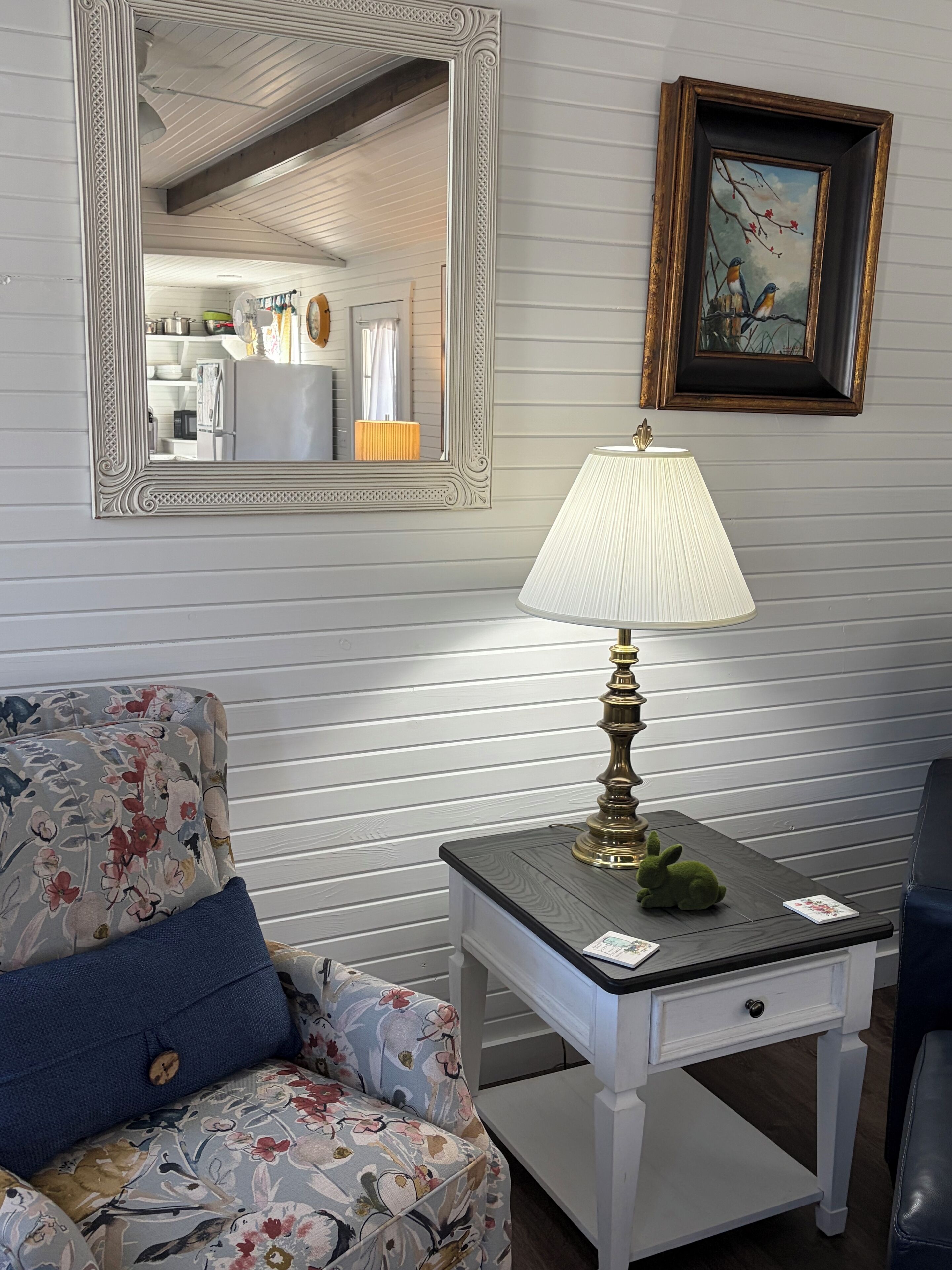 A room with a floral-patterned armchair, a small table with a lamp, a framed picture, and a mirror reflecting a kitchen area.