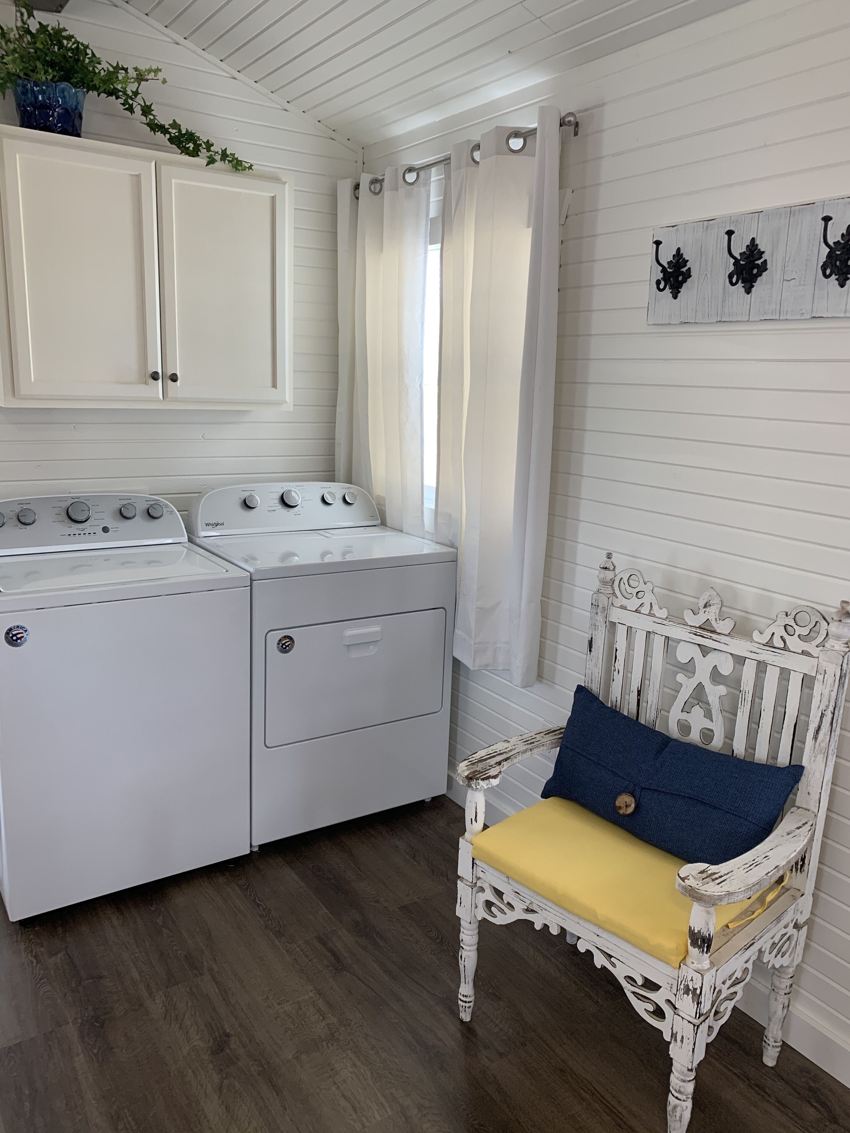 A laundry room with a white washing machine and dryer, a wooden bench with a cushion, and a window with curtains.