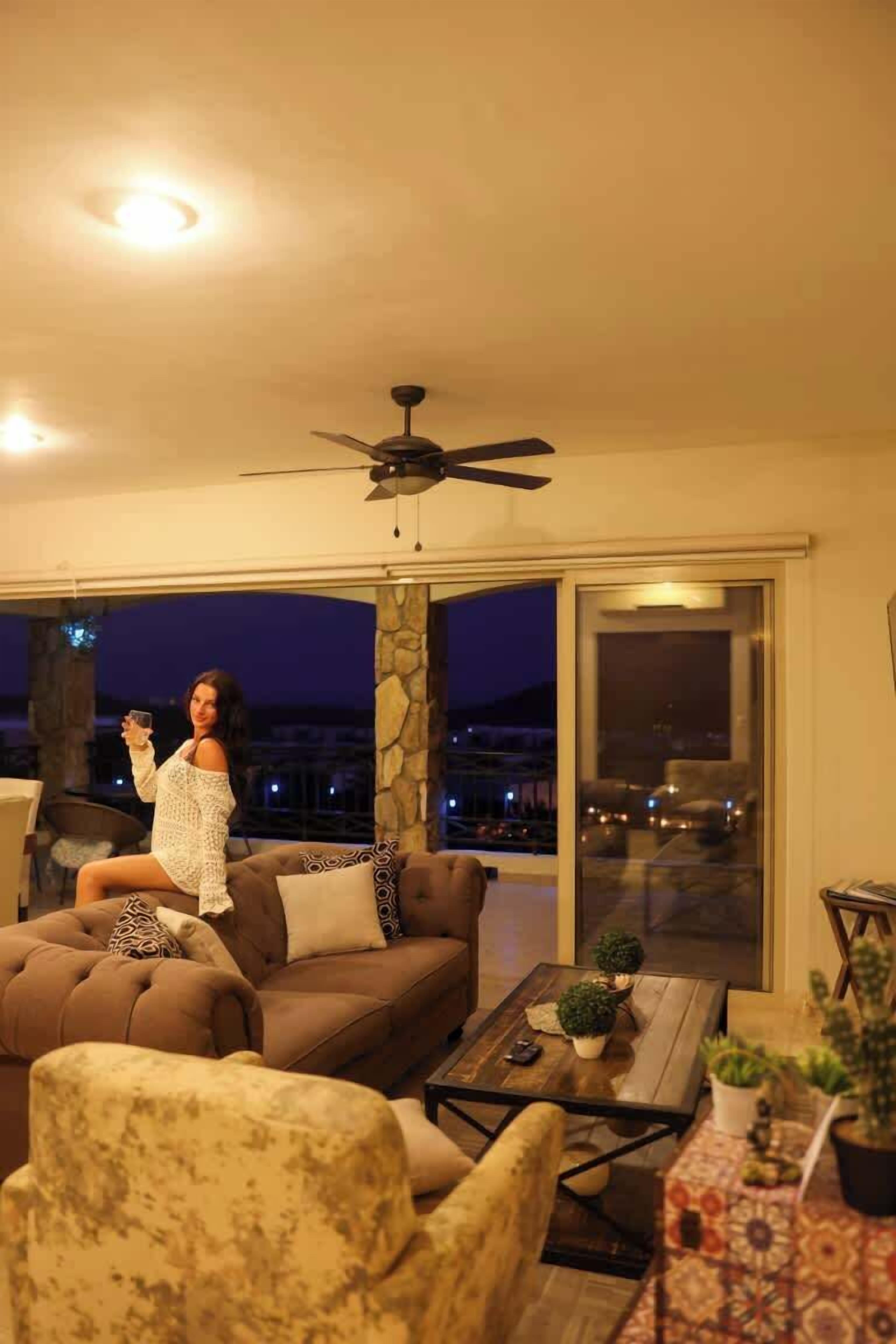 A cozy living room with a brown sofa, a patterned armchair, and a coffee table with plants. A ceiling fan is visible above the sofa.
