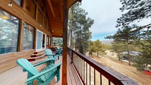 A wooden deck with Adirondack chairs, a wooden railing, and a view of a forest and mountains.
