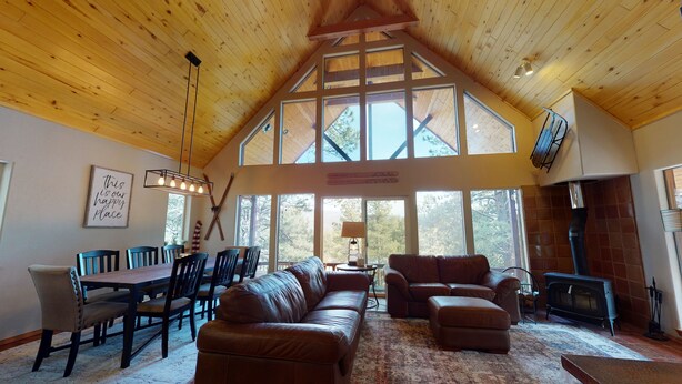 A spacious living room with a high ceiling, wooden beams, a fireplace, and large windows offering a view of the outdoors.