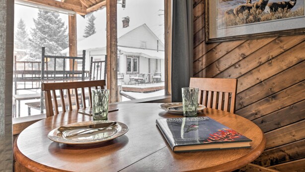 Uma mesa de jantar de madeira com um livro e copos, um quadro na parede e vista para uma casa pela janela.