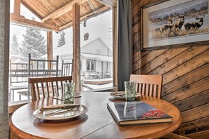 A wooden dining table with a book and glasses, a framed picture on the wall, and a view of a house through a window. - Cozy Log Cabin Escape In the Heart of Creede! (Creede)