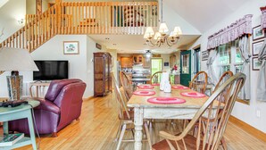 Zona pranzo con tavolo in legno, sottopiatti rossi e sedie in legno. Una scala con ringhiere in legno conduce a un piano superiore.