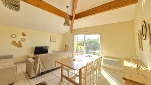 A room with a bed, a TV, a dining table with chairs, a window with a view of greenery, and wooden beams on the ceiling. - Beautiful twin villa of 100m² for 6 people (BISCARROSSE)