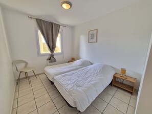 A simple, clean room with a bed, a chair, a window with curtains, and a small bedside table with a lamp.