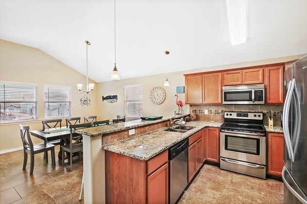 A kitchen with granite countertops, stainless steel appliances, wooden cabinets, and a dining area with a table and chairs. - Flock On-Pet friendly beach house close to the beach, golf cart zone (Port Aransas)