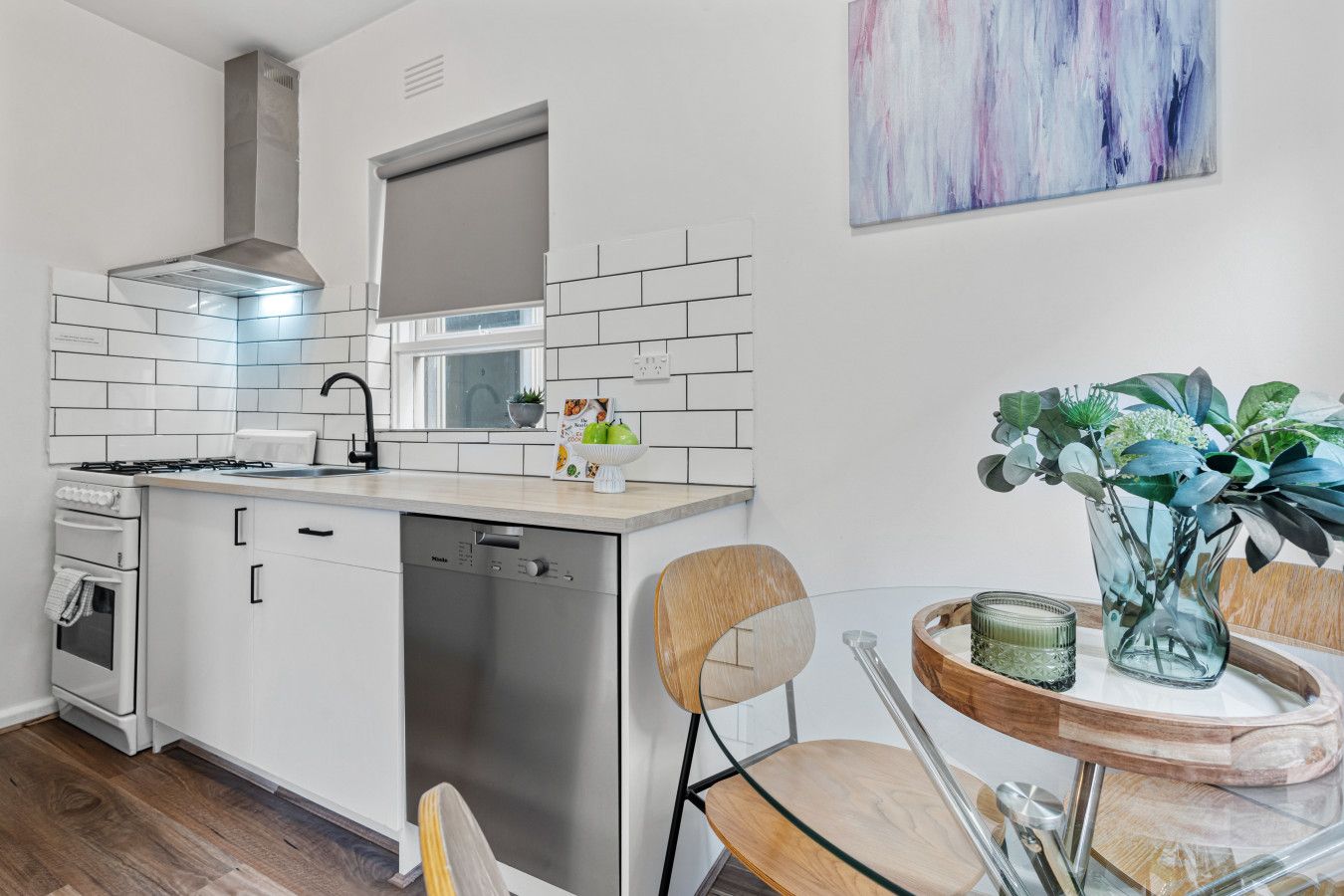 A modern kitchen with white cabinets, a stainless steel range hood, and a dishwasher. There's a dining area with a glass table and chairs, and a vase with green plants.