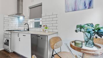 A modern kitchen with white cabinets, a stainless steel range hood, and a dishwasher. There's a dining area with a glass table and chairs, and a vase with green plants.