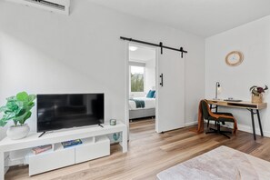 A modern living room with a flat-screen TV, a white entertainment unit, a wooden desk with a chair, and a sliding door leading to a bedroom.
