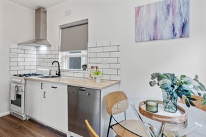 Una cocina moderna con armarios blancos, una campana extractora de acero inoxidable y un lavavajillas. Hay una ventana sobre el fregadero, una pequeña planta en maceta y un jarrón decorativo con follaje.