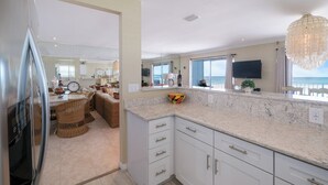 A modern kitchen with white cabinets, a marble countertop, and a large window overlooking the beach.