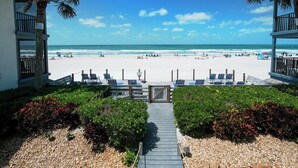 A beach view from a resort with a wooden walkway leading to the beach, lounge chairs, and a clear blue sky.