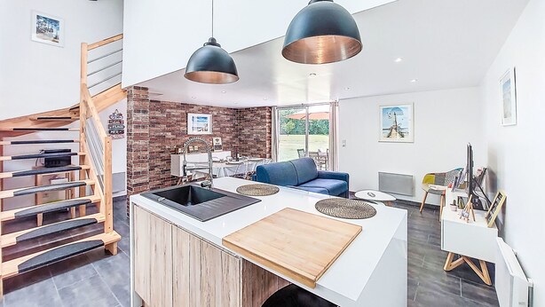 Une cuisine moderne avec un îlot central, un escalier en bois et un coin repas agrémenté d’un canapé bleu.