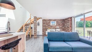Un salon moderne avec un canapé bleu, un escalier en bois et un coin cuisine doté d’un luminaire suspendu.