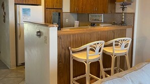 A compact kitchen with wooden cabinets, a refrigerator, a sink, and a bar area with two white bar stools. - Hawaiian Prinicess one bedroom
1990/NUC-2240 (Waianae)