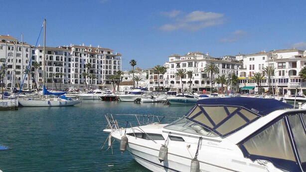 A marina with several boats docked, surrounded by multi-story buildings with balconies and palm trees. - One bedroom on the ground floor (Manilva)