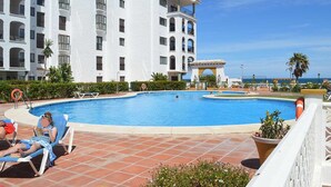 A swimming pool area with lounge chairs, a building with balconies, and a clear sky. - One bedroom on the ground floor (Manilva)