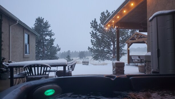 A hot tub with a glowing light, surrounded by snow-covered furniture and a patio area with string lights.
