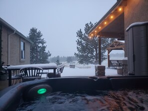 Un bain à remous illuminé, entouré de meubles recouverts de neige et d’un espace patio décoré de guirlandes lumineuses.