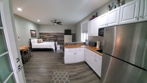 A modern kitchen with white cabinets, a stainless steel refrigerator, and a wooden countertop. There is a bed with a wooden headboard in the background.