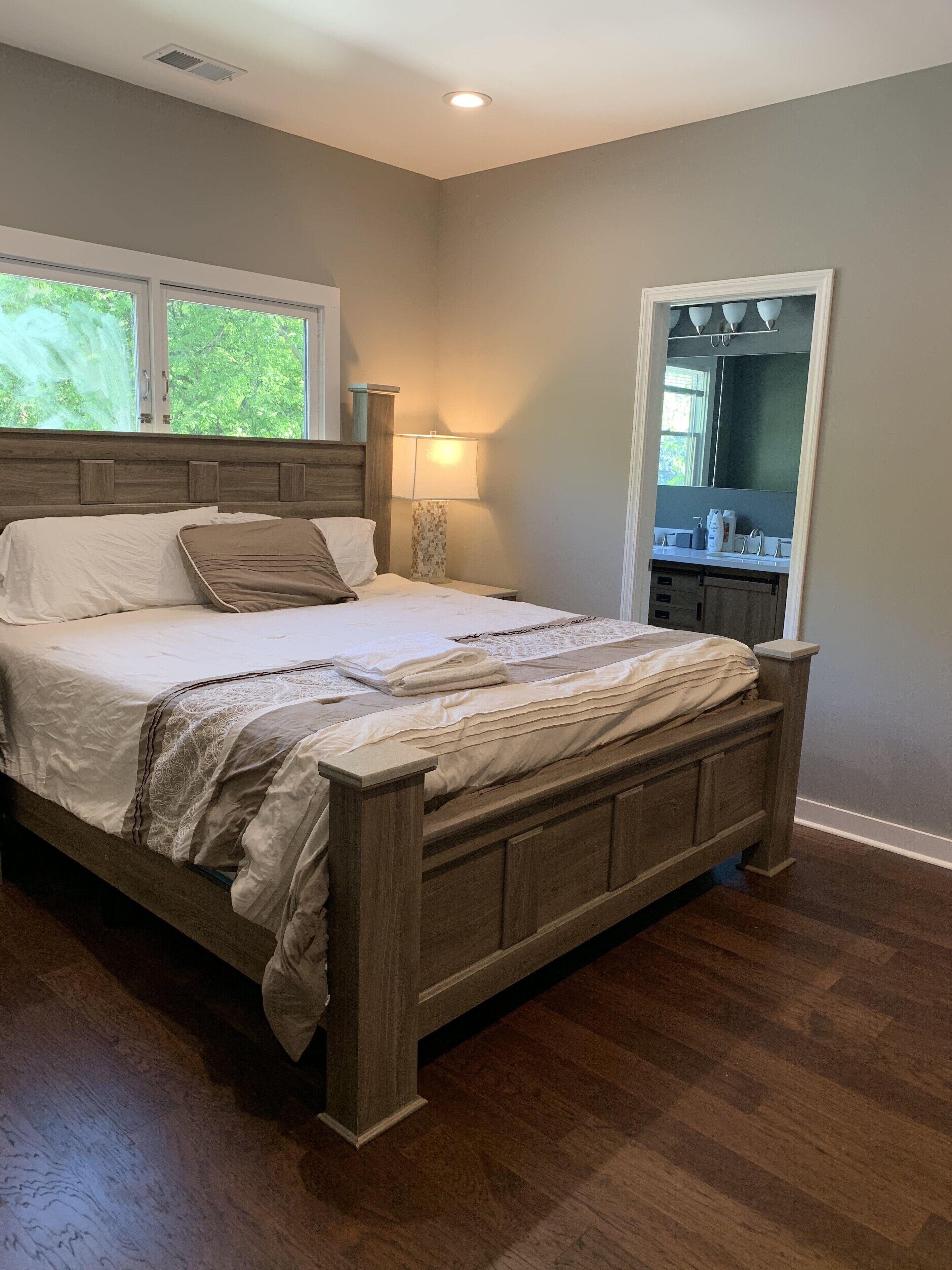 A bedroom with a wooden bed, a nightstand with a lamp, a window with a view of trees, and a bathroom with a mirror and sink.