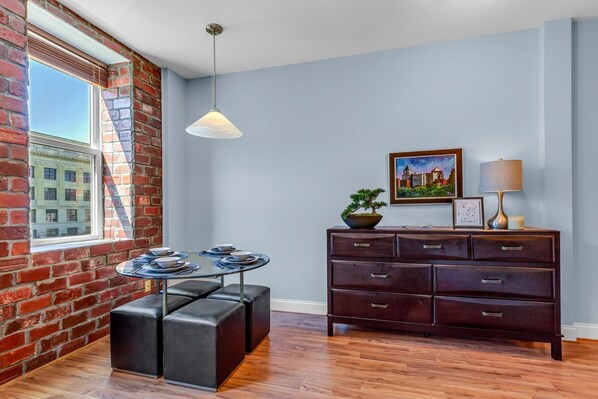 Une pièce avec un mur en briques, une table ronde entourée de tabourets, une commode en bois sombre, une lampe suspendue et un tableau encadré au mur.