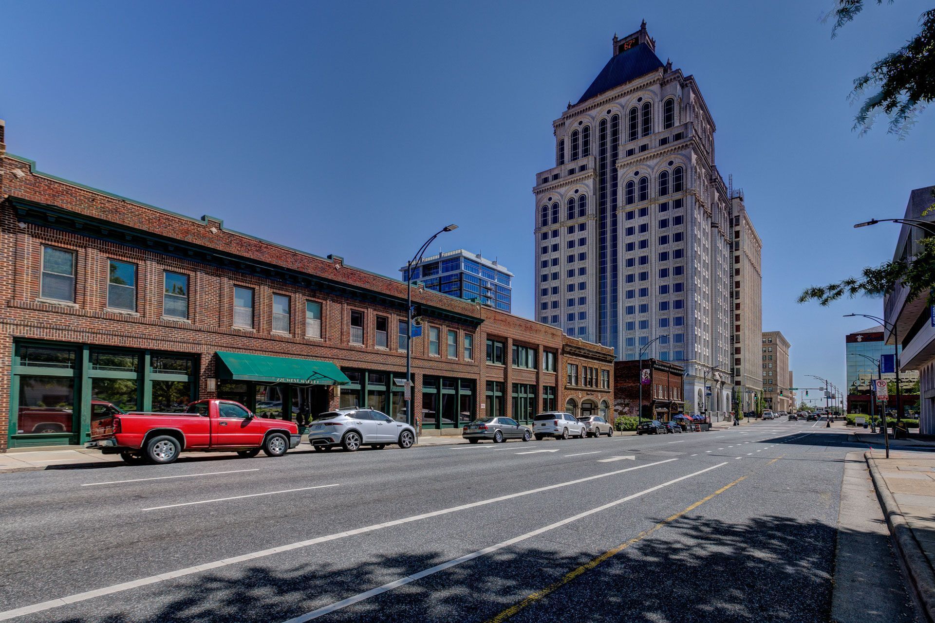 A street with a row of brick buildings, a tall building in the background, and cars parked along the side.