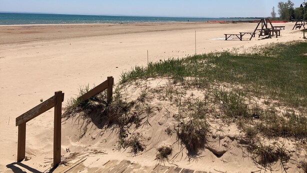 A beach view with a wooden deck, a sandy area with grass, and a clear sky. - Lakeside condo. (Oscoda)