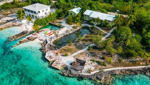 An aerial view of a coastal property with a large house, a smaller building, and a dock extending into turquoise waters.
