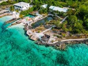 An aerial view of a coastal property with a large house, a smaller building, and a dock extending into turquoise waters.