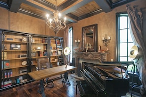 Una habitación con un piano de cola, una mesa de madera, una estantería, una araña y un espejo grande.