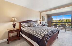 Une chambre d’hôtel avec un grand lit, une table de chevet, une lampe, un bureau avec une chaise et une vue sur l’extérieur grâce à une grande fenêtre.