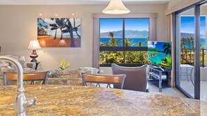 A hotel room with a marble kitchen island, a dining area with a table and chairs, a sofa, a TV, and a large window with a view of the ocean and mountains.