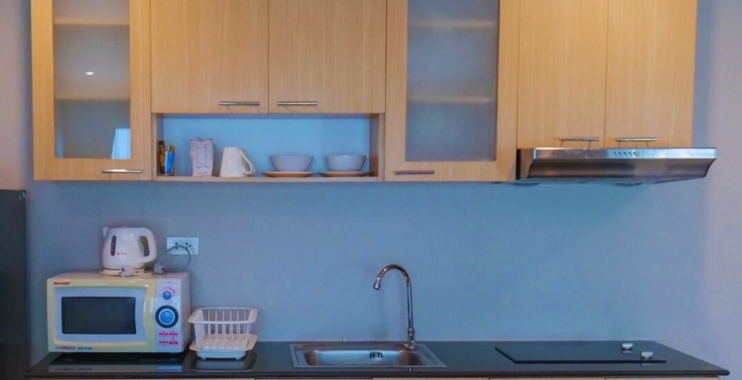 A compact kitchenette with wooden cabinets, a microwave, a kettle, a sink, and a stainless steel range hood.