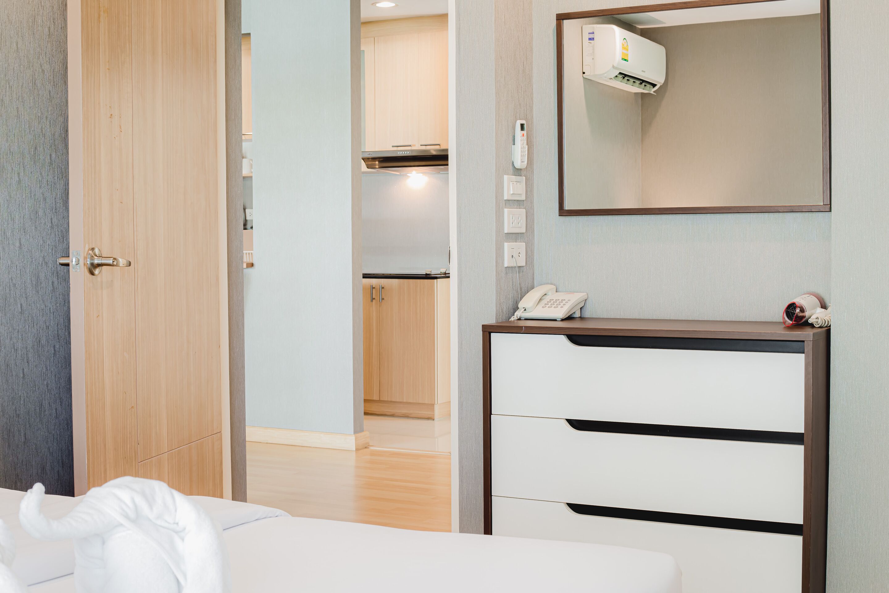 Habitación de hotel con puerta de madera, cama blanca, mesita de noche con teléfono y espejo.