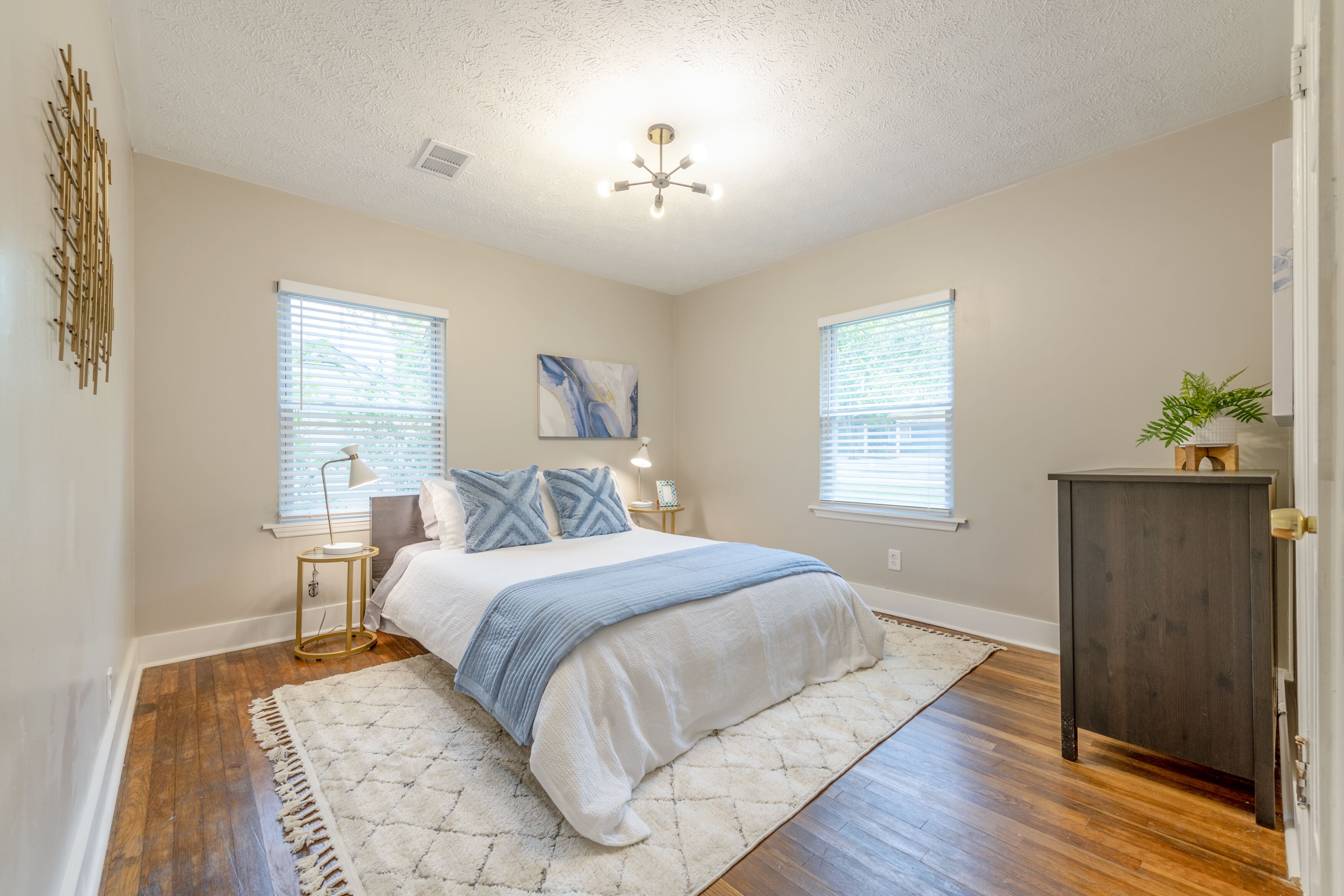 A bedroom with a bed, bedside lamps, a nightstand, a painting, and a plant.