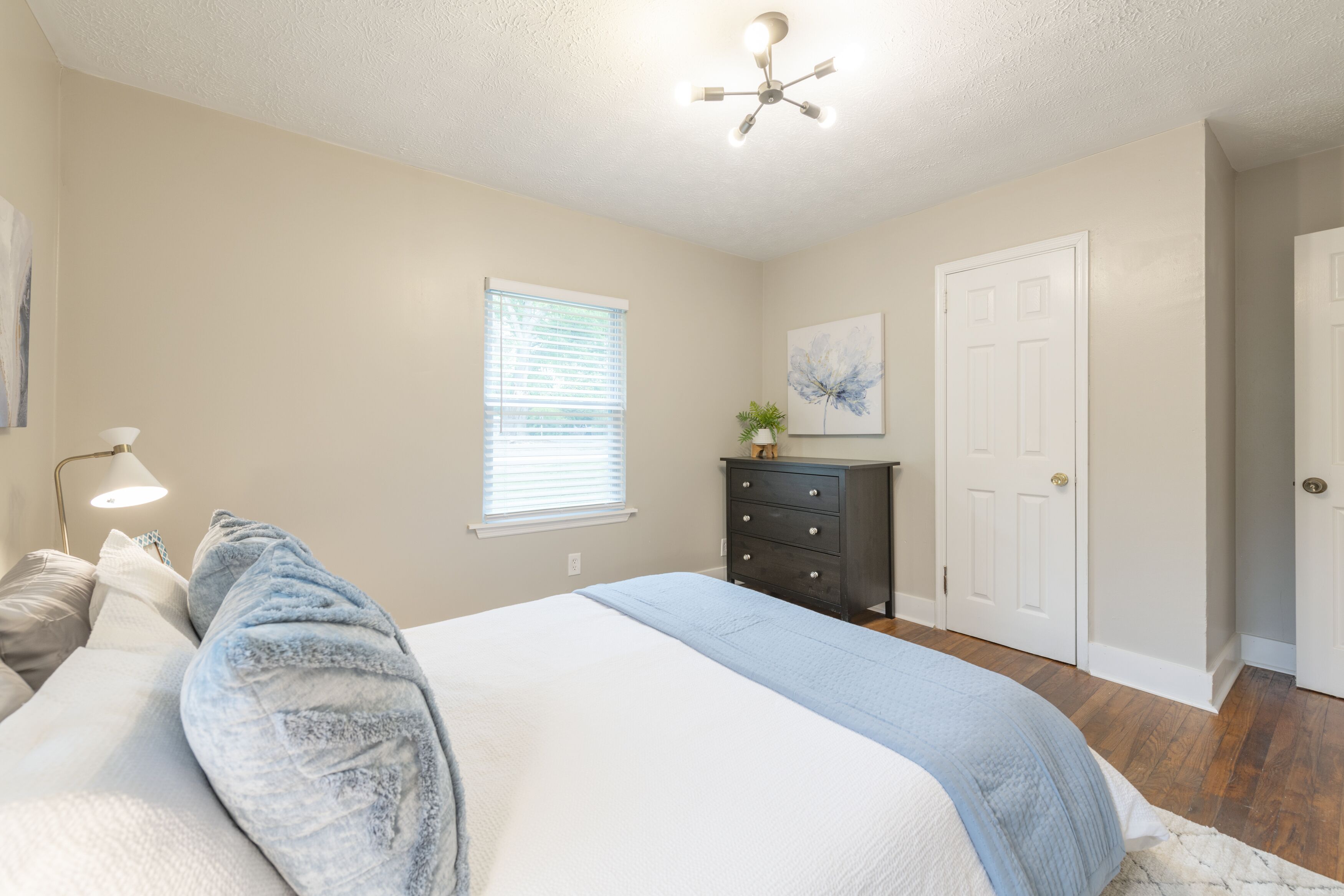 A bedroom with a bed, a dresser, a ceiling fan, a window with blinds, and a door.