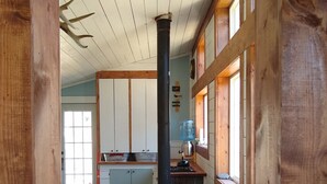 A rustic cabin interior with a wooden interior, a black stove, and a view of a kitchen area.