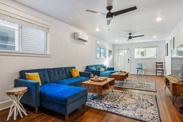 A spacious living room with a blue sofa, a wooden coffee table, and a patterned rug. There are ceiling fans, a TV, and a door leading to another room. - Mid-City NOLA Getaway, Great for Groups! (New Orleans)