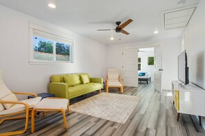 A modern living room with a green sofa, two armchairs, a ceiling fan, a flat-screen TV, and a large window with a view of trees. - Mid-City NOLA Getaway, Great for Groups! (New Orleans)