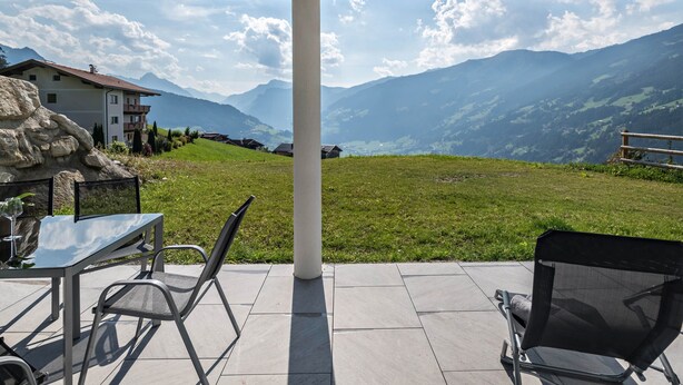 Eine Terrasse mit Tisch und Stühlen, Blick auf Berge und ein Gebäude im Hintergrund.