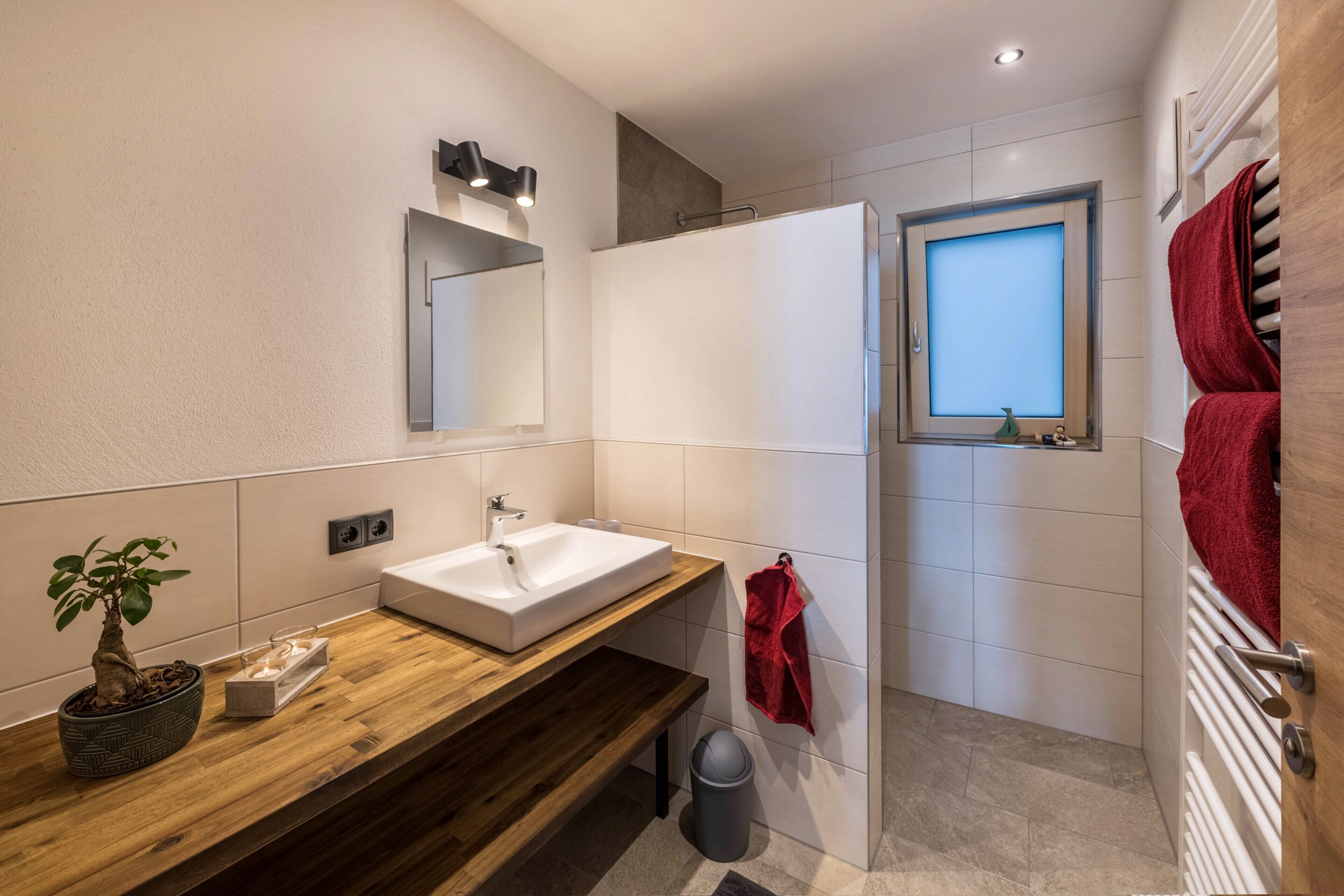 Ein modernes Badezimmer mit einem Holzkosmetikschrank, einem weißen Waschbecken, einem Spiegel, einem roten Handtuch und einem Fenster.