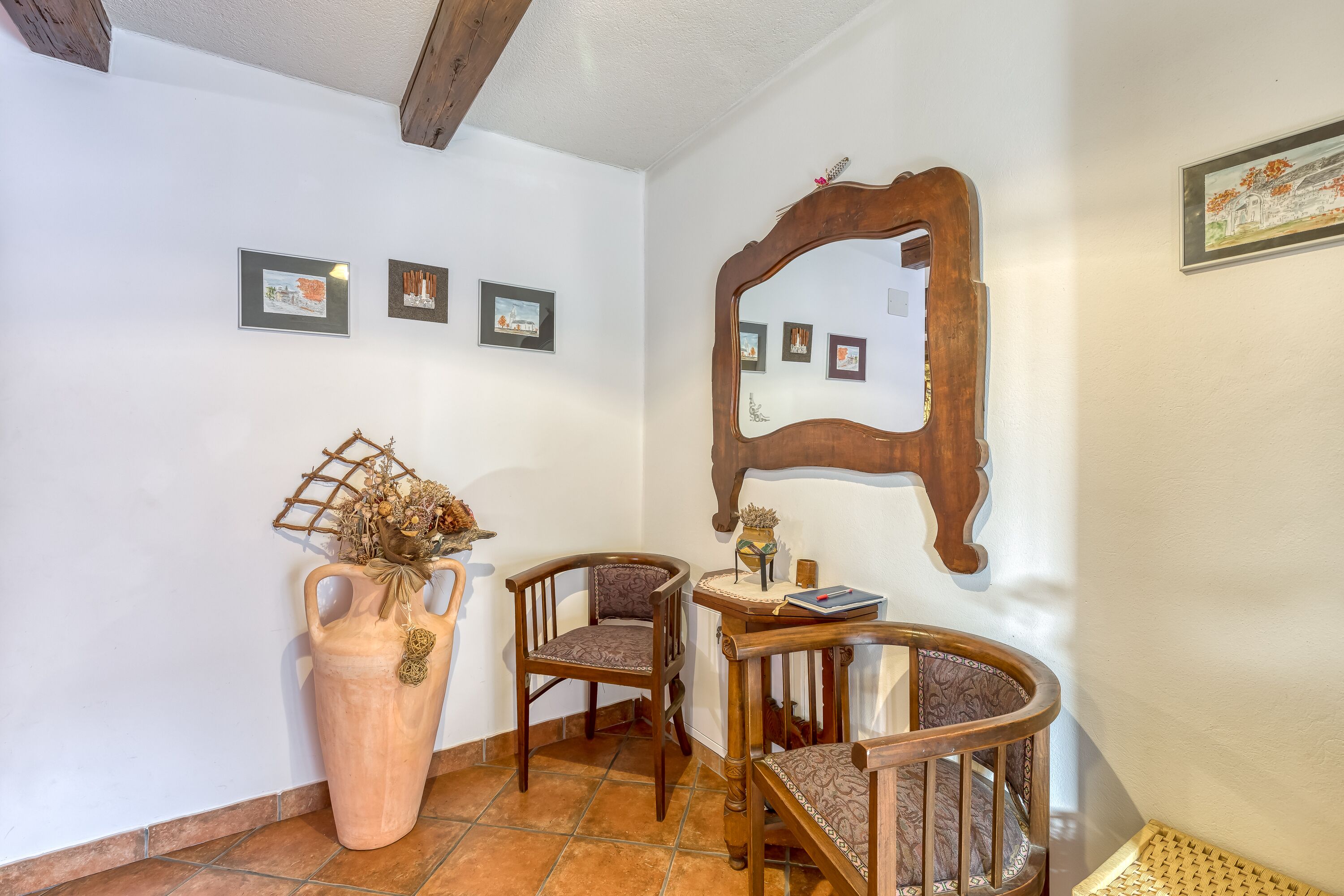 A corner with a large vase, two wooden chairs, a small table, and a mirror on the wall.