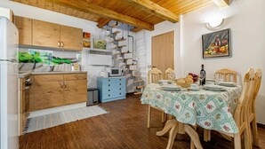 Une cuisine avec des meubles de rangement en bois, une commode bleue, un coin repas avec une table dressée pour quatre personnes et un escalier menant à l’étage supérieur.