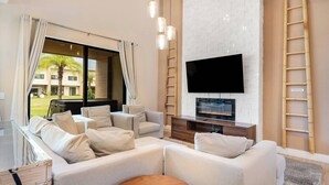 A modern living room with a large sectional sofa, a flat-screen TV mounted on a white brick wall, and a wooden ladder leaning against the wall. - 7458 Modern new home 3 suites jacuzzi 8 min Disney (Kissimmee)