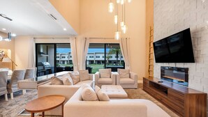 A modern living room with a flat-screen TV, beige sofas, a wooden coffee table, and a large window offering a view of a building and palm trees. - 7458 Modern new home 3 suites jacuzzi 8 min Disney (Kissimmee)