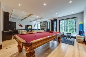 Une table de billard dans une pièce dotée d’une cheminée en pierre, de grandes fenêtres et d’une vue sur une forêt.