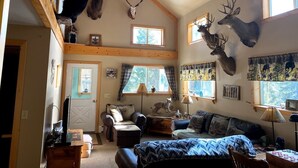 Interior acollidor d'una cabana amb un llit d'altalç, una xemeneia i uns corrons de cérvol penjats a les parets.
