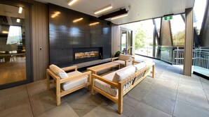 A modern lounge area with a fireplace, wooden furniture, and large windows offering views of the outdoors. - Sudima Queenstown Five Mile (Queenstown)